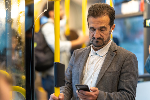 Mature businessman using phone and listening to music while standing in bus.