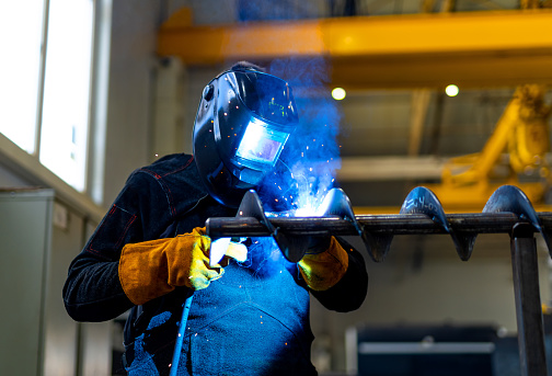 A welder welds metal into his workshop. Blue welding sparks. Gas combustion and blue smoke. Small welding workshop. Welding juncture of metal construction