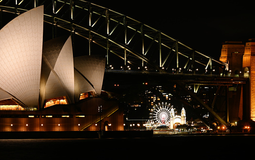 A view across the harbour of Sydney's two most iconic landmarks, the Opera House and Harbour Bridge, lit up at night. The Ferris Wheel of the Luna World amusement park can be seen beneath the bridge.