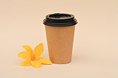 A cardboard cup for coffee, suitable for reuse, next to a yellow lily flower on a beige background, zero waste, careful attitude to nature.