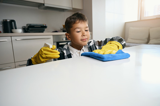 Teenager in a plaid shirt and protective gloves helps around the house, he washes the kitchen surface