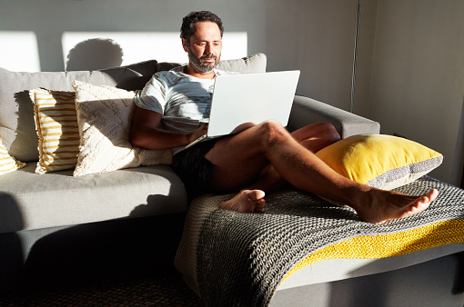 portrait mature man working on his laptop relaxed in shorts and t-shirt on the couch from home