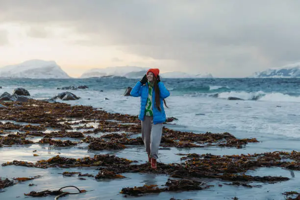 Happy female with long hair and inblue jacket walking at the scenic beach with cold ocean view in Scandinavia