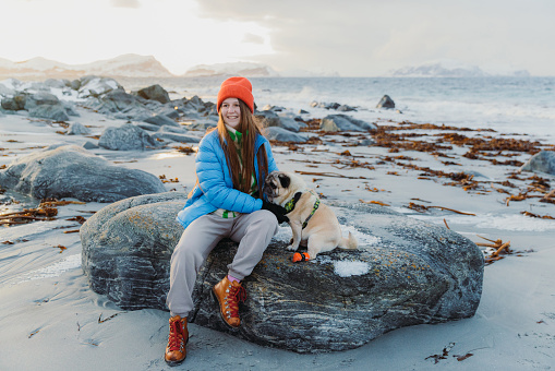 Front view of smiling female in red hat and blue jacket petting her cute pug at the beautiful beach with snowcapped island view during idyllic sunset in Scandinavia