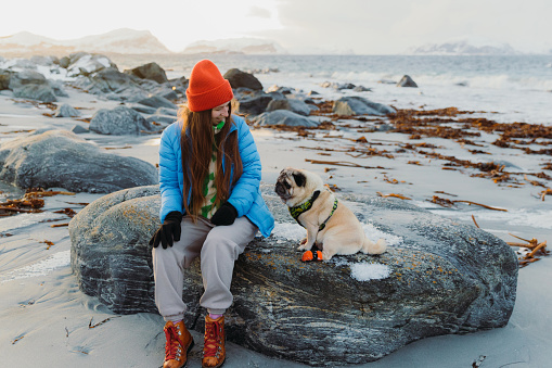 Front view of smiling female in red hat and blue jacket petting her cute pug at the beautiful beach with snowcapped island view during idyllic sunset in Scandinavia