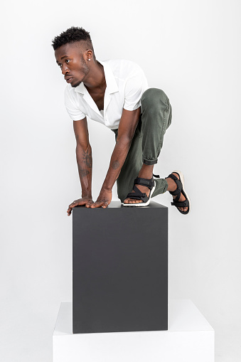 Black handsome man of African descent wearing white shirt, brown fabric pants, black summer sandals with cubes in studio.