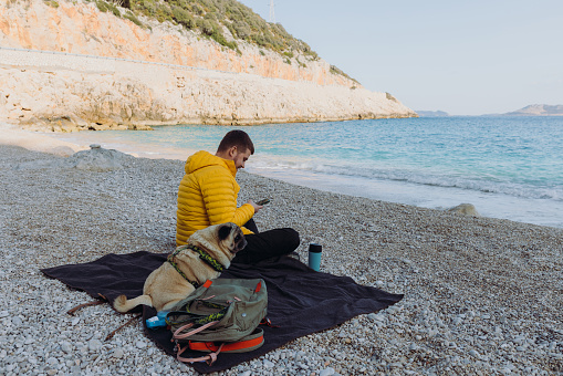 Side view of a male in yellow jacket relaxing on picnic blanket with his pug at the beach text messaging by phone with scenic sea view in Antalya province, Middle East