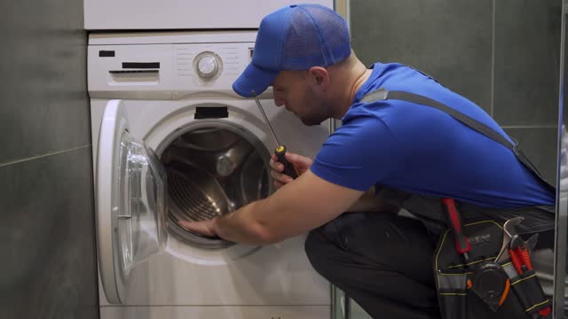 Installation or repair of a washing machine. A plumber in a blue uniform and a cap repairs a washing machine in the bathroom