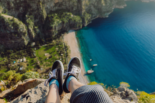 High-angle view of female's legs wearing sneakers sitting at the edge of the mountain with view of crystal blue sea at Butterfly Valley in Turkey, Middle East