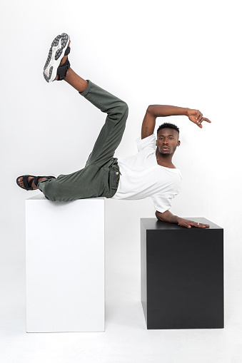 Black handsome man of African descent wearing white shirt, brown fabric pants, black summer sandals with cubes in studio.