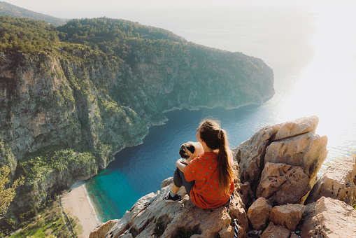 Happy female sitting on the cliff with a dog enjoying the bright sunset above the Butterfly Valley in Mediterranean Turkey