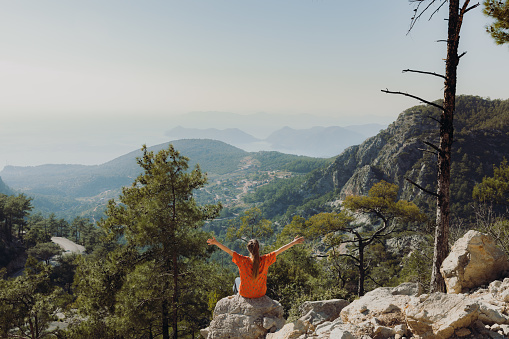 A woman traveler enjoying mountain trip in Mediterranean Turkey and sitting on the viewpoint raising hands with dramatic view of raw nature during bright sunset