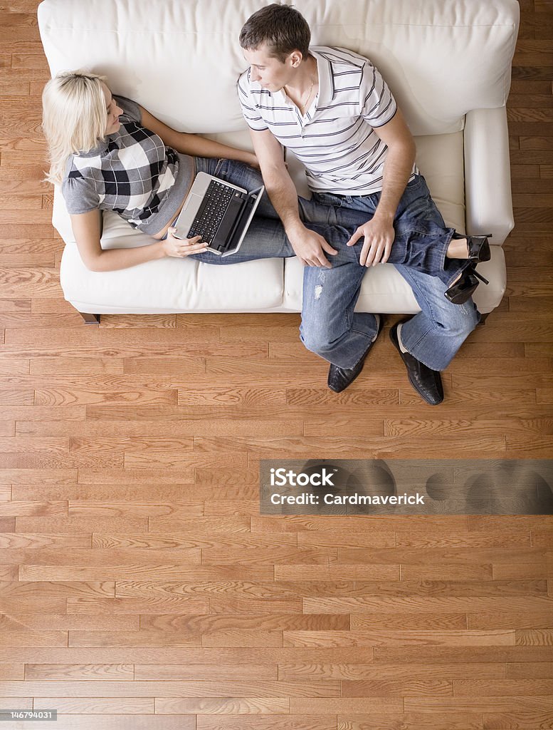 Overhead View of Couple Relaxing on Couch Full length overhead view of couple relaxing together on white couch, with woman using laptop and stretching out with her legs in the man's lap. Vertical format. Hardwood Floor Stock Photo