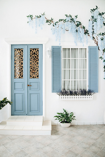 Blue door and window on the white wall of a mediterranean-style house with a flowering tree weaving along the wall. Porch of a house in Santorini.
