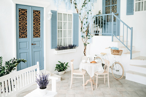 Mediterranean style white house exterior with blue door and window, flowering tree and romantic seating area. Traditional patio of Santorini.