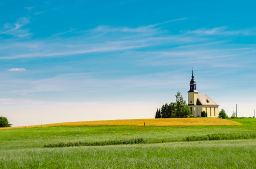 horizontal image of a quaint little white country church with a steeple sitting in a green meadow surrounded by trees under a beautiful blue sky with white clouds floating by in the summer time.
