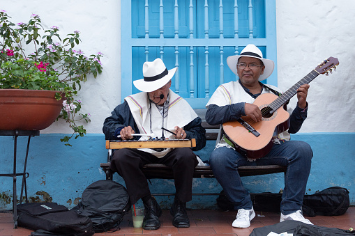 Medellin, Colombia - July 30, 2022: Two senior musicians of traditional Colombian music, one of them playing a xylophone and the other a guitar. Both are sitting on a bench outside a house.