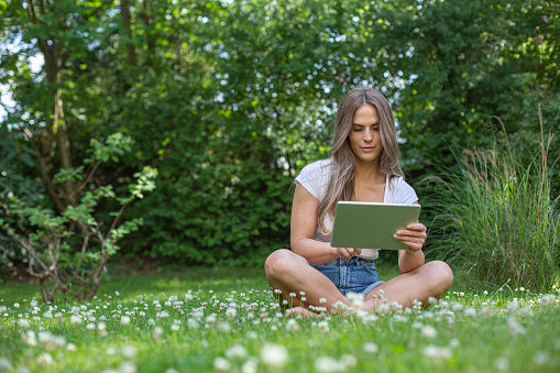 Young pretty blonde woman sitting in the garden with an tablet and reading, playing, online banking or using an app