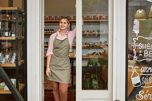 Front view of sales clerk in casual attire and apron standing at entrance to bath and beauty shop and smiling at camera.
