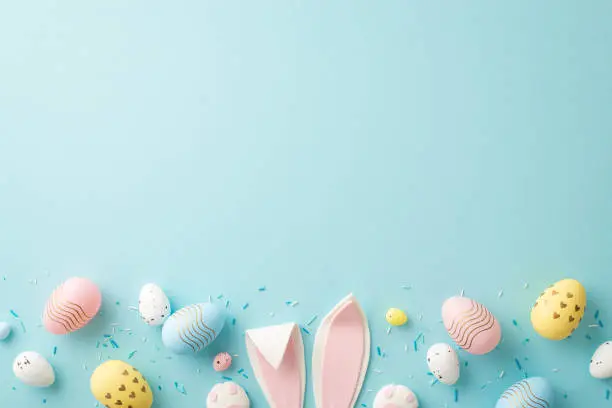 Photo of Easter concept. Top view photo of easter bunny ears colorful eggs and sprinkles on isolated light blue background with copyspace