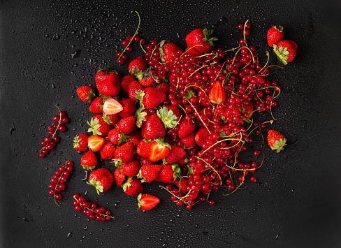 Strawberries and red currants on a dark background. Sprigs of red currant and strawberries with green tails on a black background with water drops top view.