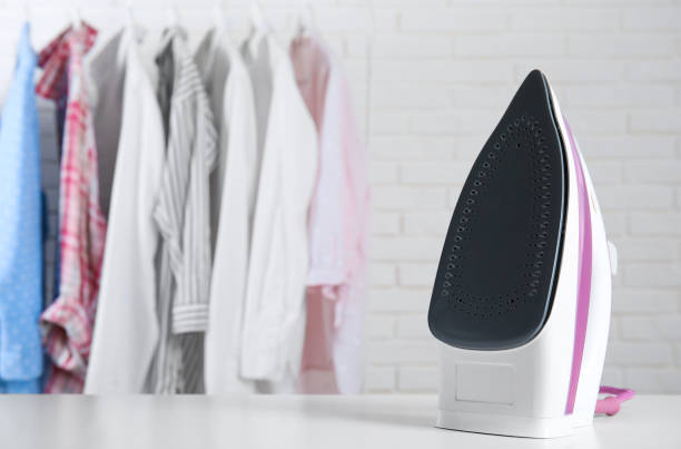 istock Modern iron on table near rack with clean clothes indoors, space for text 1467970917
