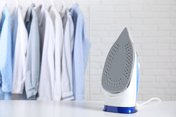 istock Modern iron on table near rack with clean clothes indoors, space for text 1467970946