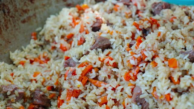 The close -up of rice with lamb is interfered with a spoon. The cauldron of pilaf is prepared on fire. The male hand in the glove closes the hot cover. Hot dish in nature, lunch is ready.