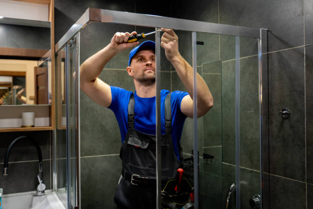 Young man in uniform repairs the shower door in the bathroom. A male repairman repairs the shower cabin stock photo