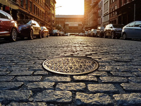 Manhole cover in an old cobblestone street lined with parked cars in the Tribeca neighborhood of New York City NYC