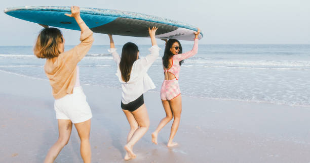 group of happy young asian girl friends carry stand up paddleboard and run on beach, smile and laugh together. asia summer vacation, sup boarding, woman travel lifestyle, or leisure activity - women paddleboard bikini surfing imagens e fotografias de stock