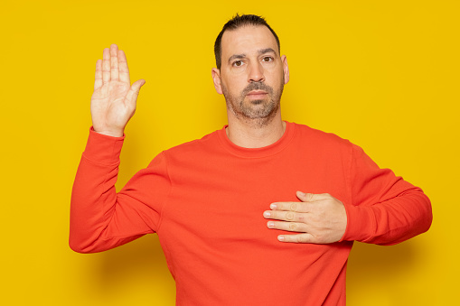 I swear to be honest. Sincere responsible handsome bearded hispanic man raising hand to take oath, promising to be honest and tell the truth, keeping hand on chest. Adult man isolated on yellow background