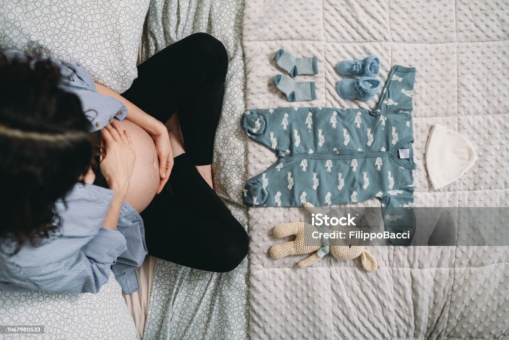 A pregnant woman is looking the clothes of her future baby on the bed A pregnant woman is looking the clothes of her future baby on the bed. She's waiting for her baby. Infant Bodysuit Stock Photo