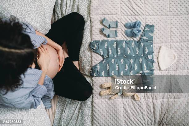 A Pregnant Woman Is Looking The Clothes Of Her Future Baby On The Bed Stock Photo - Download Image Now