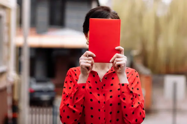 Confident smiling woman in glasses is showing red paper notepad with mock up and looking at camera outdoors on street background. Female in red polkadots blouse. Concept of advertising
