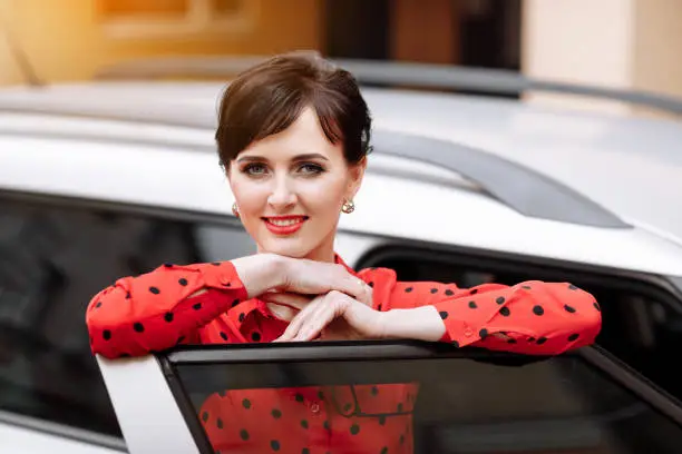 confident businesswoman looking at camera and smiling by open door car. Cheerful successful lady with red lipstick in elegant red polkadots blouse by the vehicle. Business concept.