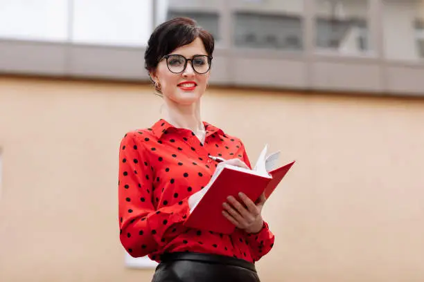 Confident businesswoman in glasses is doing notes in paper notepad by the office buildings background. Executive in red polkadots blouse is resolving work issues during the lunch break outdoors