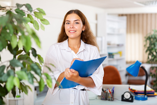 Portrait of friendly young female administrative secretary standing in office with folder of documents in hands, writing down tasks