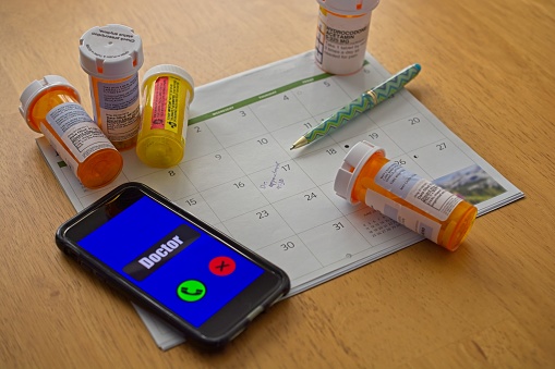 Cell phone call to doctor with prescription drug bottles strewn across calendar highlighting appointment reminder on wooden table. High angle view.