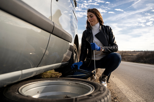 A young woman is changing a flat tire on her car on the road.