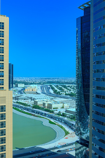 It is early morning in the Arabian City of Dubai in the United Arab Emirates. The view from a tower in the Jumeirah Lake Towers (JLT) area, looking towards a residential area. The desert sand that can be seen here and there is how the area should really look like! In the foreground is a small lake. There is traffic on the road below. Photo shot in the morning sunlight; vertical format. Copy space. No people.