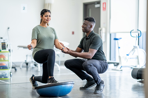 A male physiotherapist of African decent, works with a young female client in a gym.  The client is dressed comfortably as they work in the gym with a half ball on strength and balance.