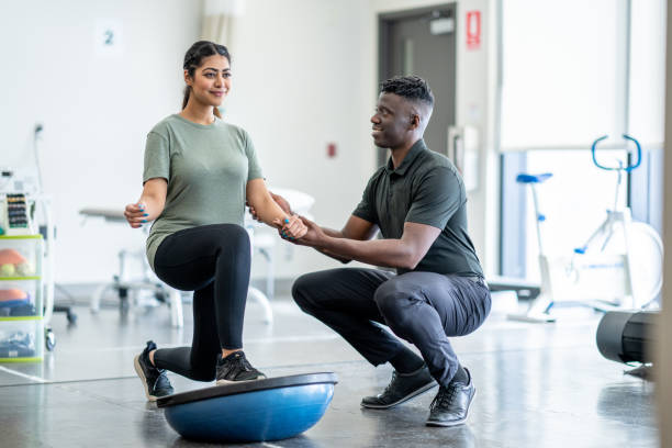 Working on Balance A male physiotherapist of African decent, works with a young female client in a gym.  The client is dressed comfortably as they work in the gym with a half ball on strength and balance. sports medicine stock pictures, royalty-free photos & images