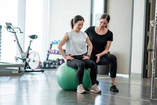 A young female works with her physiotherapist in a gym during an appointment.  She is dressed comfortably and sitting on a yoga ball as they work through exercises to strengthen her core.