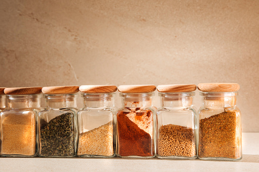 A group of seasonings in glass jars on a light stone background with shadows. Paprika, herbs, mustard, garlic, selective focus