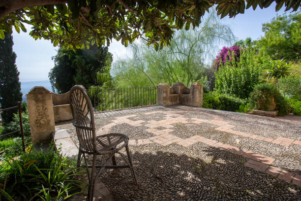 Fantastic terraced garden in the old villa Casa Cuseni in Taormina: gardening in Sicily in southern Italy.  Unreal Caesalpinia brasiliensis tree and purple ,bougainvillea purple blossoms Wooden chair and concrete floor with small stones on the terrace (garden) in the villa.