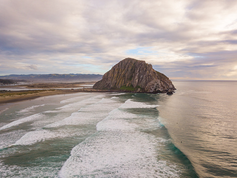 Aerial views of Morro Rock, taken with a drone in a public park, at Morro Bay State Park. High angle views taken of a large rock in coast of california with surfers riding waves.