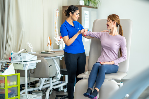 A female physiotherapist works with a client as she checks her range of motion and they discuss her limitations.  The therapist is dressed professionally as she talks with her client and they assess her movement together.