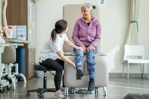 A female physiotherapist works with a client as she checks her range of motion and they discuss her limitations.  The therapist is dressed professionally as she talks with her client and they assess her movement together.