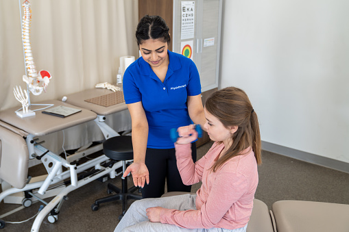 A young girl sits up on an assessment table as she works with a physiotherapist during an appointment.  The therapist is using a small dumbbell to help her client gain strength and attain her therapy goals.
